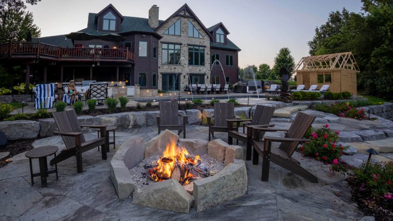 Custom landscape design with stone fire pit, chairs, and luxury home. Backyard oasis with pool, landscaping, and outdoor living.