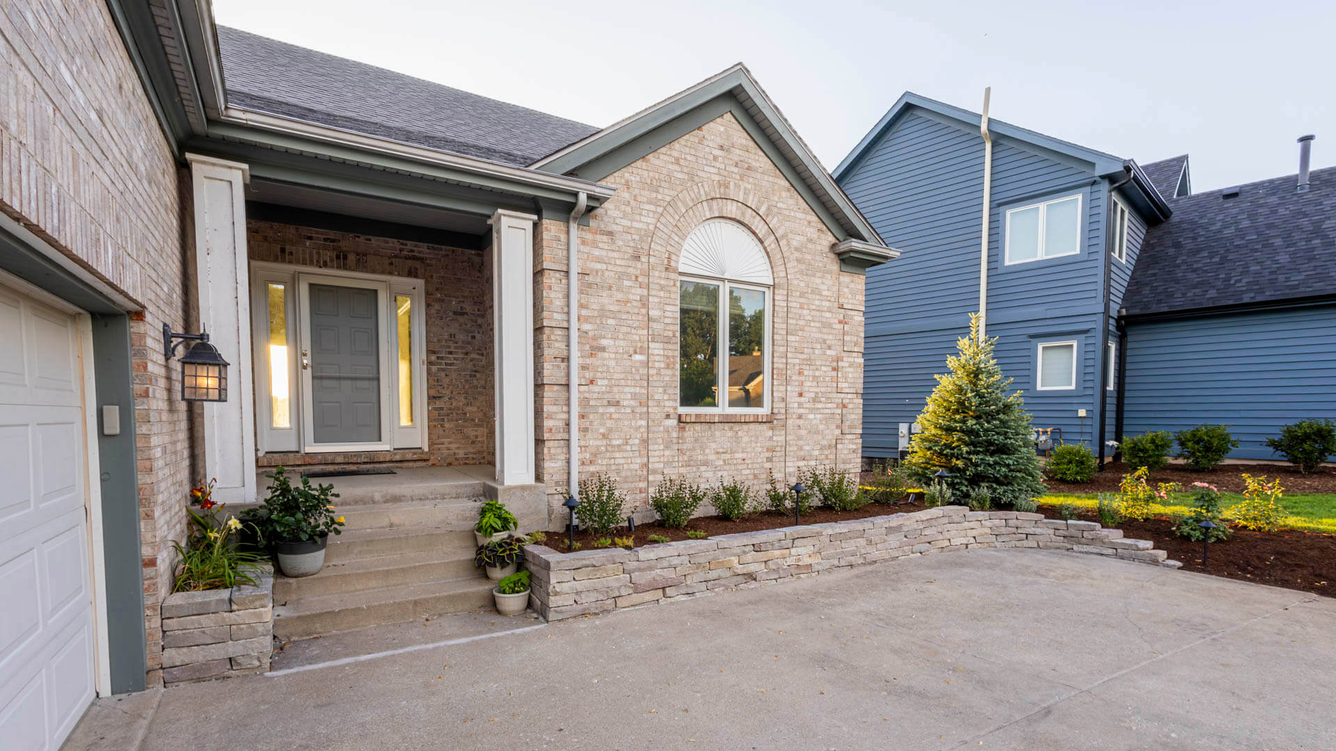 Brick home exterior with landscaping. Stone retaining wall and concrete driveway. Blue house in background.