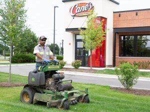 Commercial lawn mowing: Man operating a John Deere mower on a green lawn near a Raising Cane's restaurant.