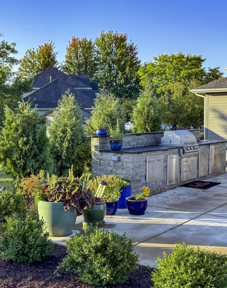 Custom outdoor kitchen with grill, landscaping, and stone countertop. Backyard design with lush greenery.