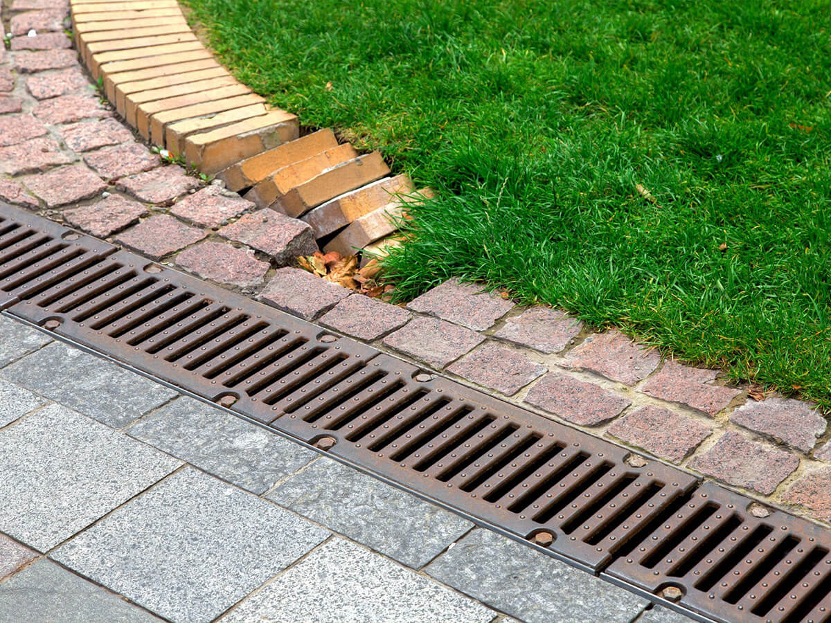 Metal trench drain installed between a paved walkway and a landscaped lawn with a curved brick border.