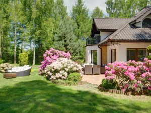 Blooming pink and white rhododendrons surround a brick patio and a house with a balcony in a lush residential garden.