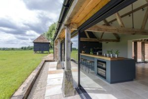 Modern home open-air kitchen and backyard. Island with stools, wooden beams, and a view of the green field.